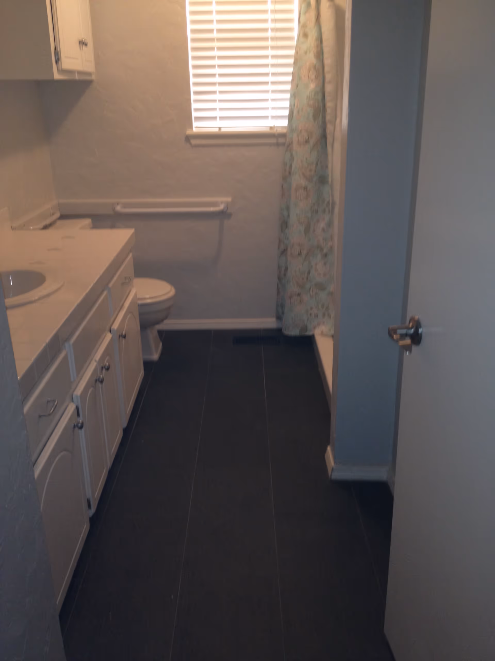 A bathroom with a white vanity and sink on the left, a toilet next to the vanity, a window with blinds above the toilet, and a shower with a floral-patterned curtain on the right side.