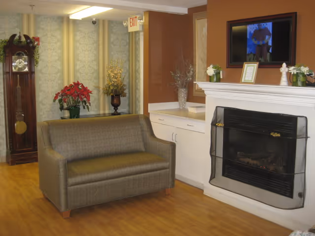 Small sitting area in a senior living facility featuring a loveseat, a fireplace with a framed TV above, a grandfather clock, and decorative plants.