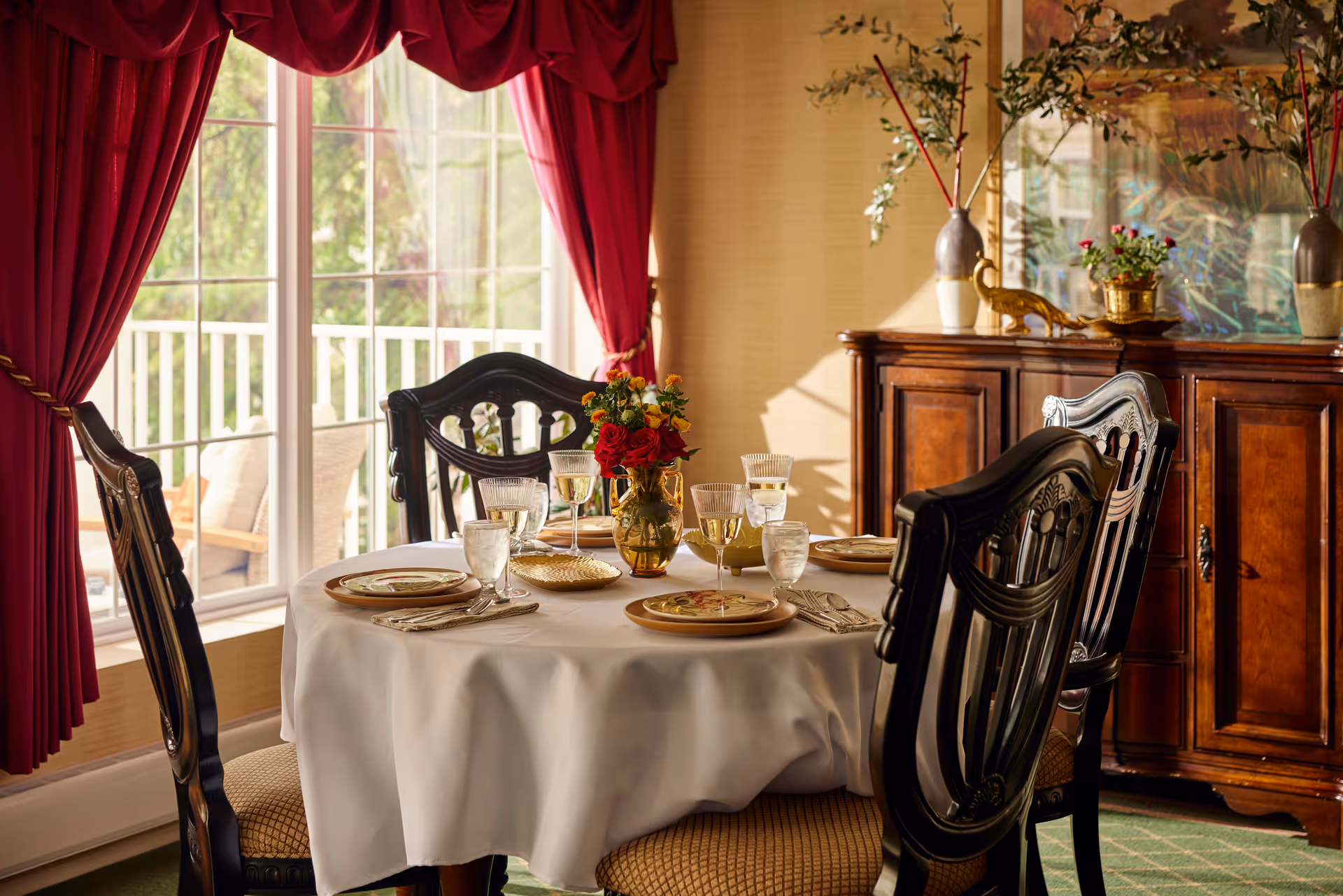 A dining room with a round table covered with a white tablecloth, set with plates, glasses, and silverware. Four dark wooden chairs with cushioned seats surround the table. A vase with red and yellow flowers is in the center of the table. Behind the table is a wooden sideboard with decorative vases and a framed artwork on the wall. Large windows with red curtains allow natural light to fill the room.