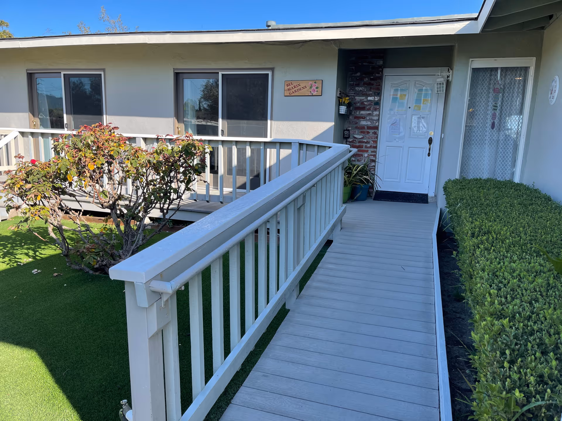 Exterior walkway ramp with white railing leading to a ground-floor apartment door, sliding windows, and shrubs.