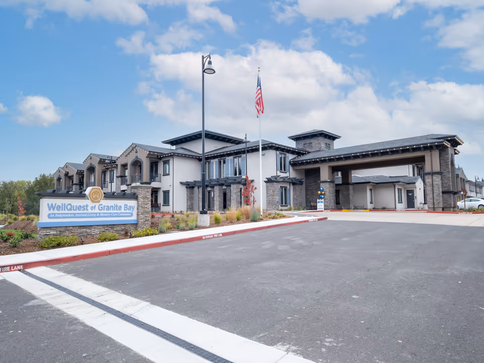 Front exterior of the WellQuest of Granite Bay senior living community showing the entrance canopy, sign, landscaping, and an American flag.
