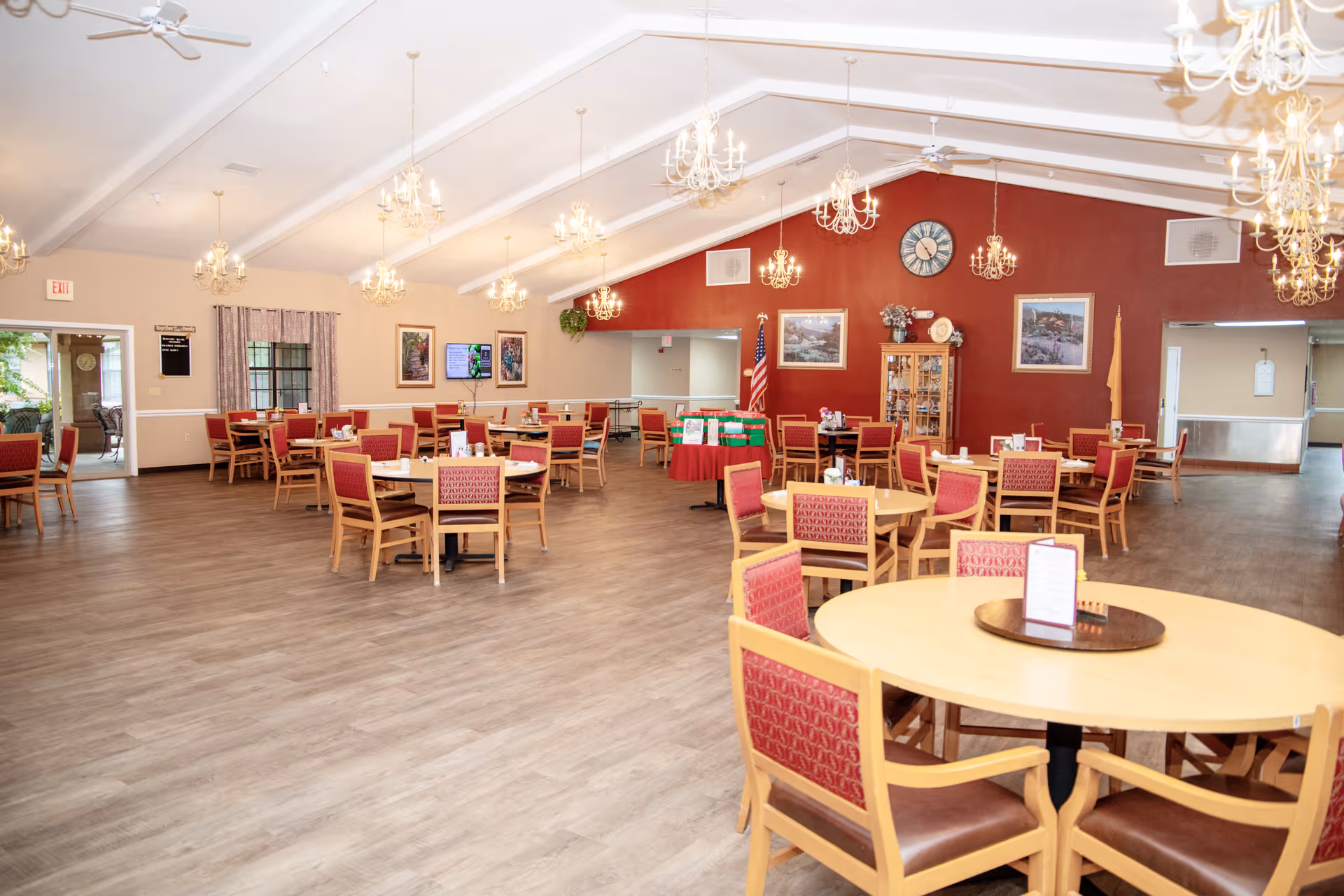 A spacious dining room with multiple round and square wooden tables surrounded by red cushioned chairs. The room features a high vaulted ceiling with several chandeliers, a red accent wall with framed pictures, a clock, and an American flag. The floor is wood laminate, and there are large windows and doorways leading to other areas.