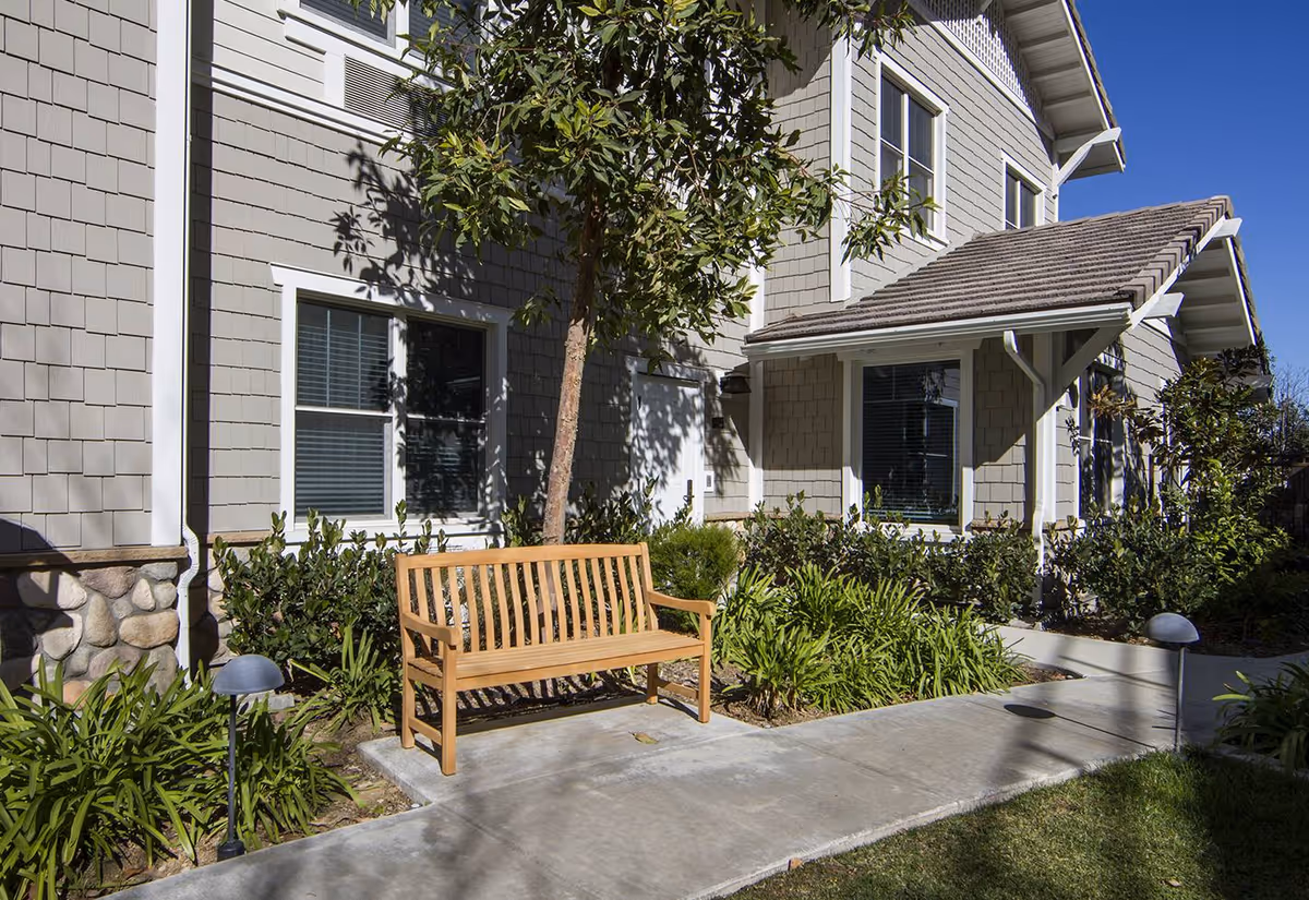 Wooden bench and landscaped walkway in front of a light-gray two-story building under a clear blue sky.