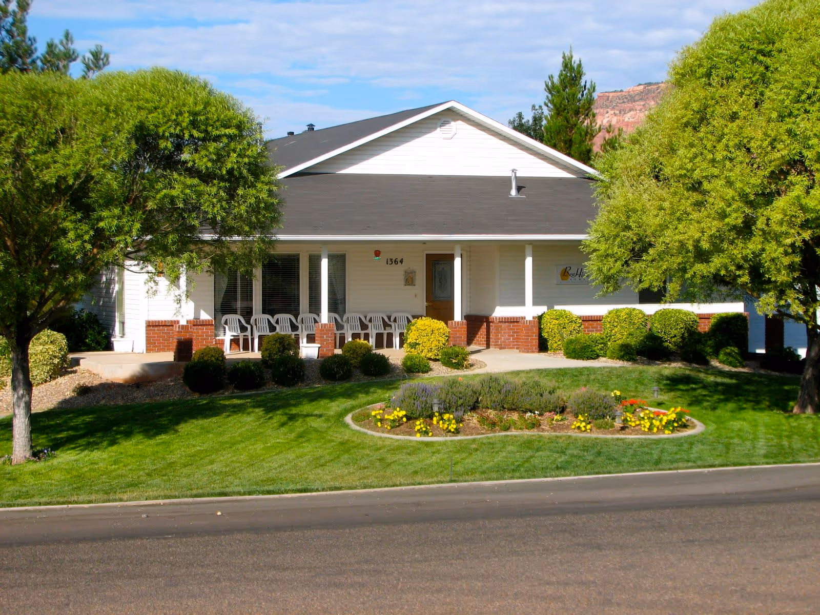 Front exterior view of a single-story white building with a dark roof, a porch with several white chairs, surrounded by green trees, bushes, and a flower bed in the lawn. The building has the number 1364 and a sign that reads Beehive Homes.