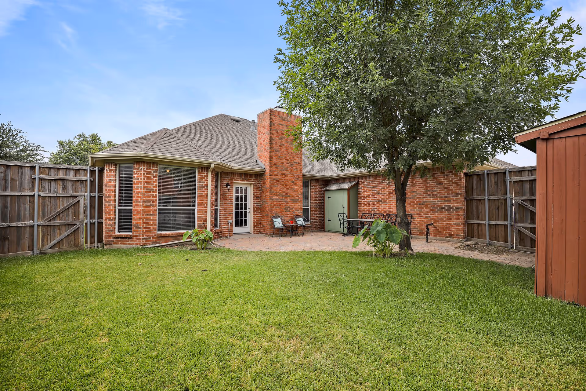 Backyard of a brick house with a green lawn, a tree, a wooden fence, a small patio area with chairs and a table, and a red garden shed on the right side.