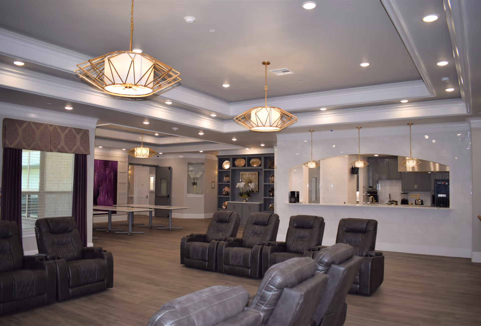 A spacious, well-lit common area with multiple dark leather recliner chairs arranged in rows facing a counter with pendant lights hanging above. The room features a modern ceiling with recessed lighting and decorative hanging lamps. In the background, there is a kitchen area visible through a large pass-through window, and a table with chairs near a window with curtains.