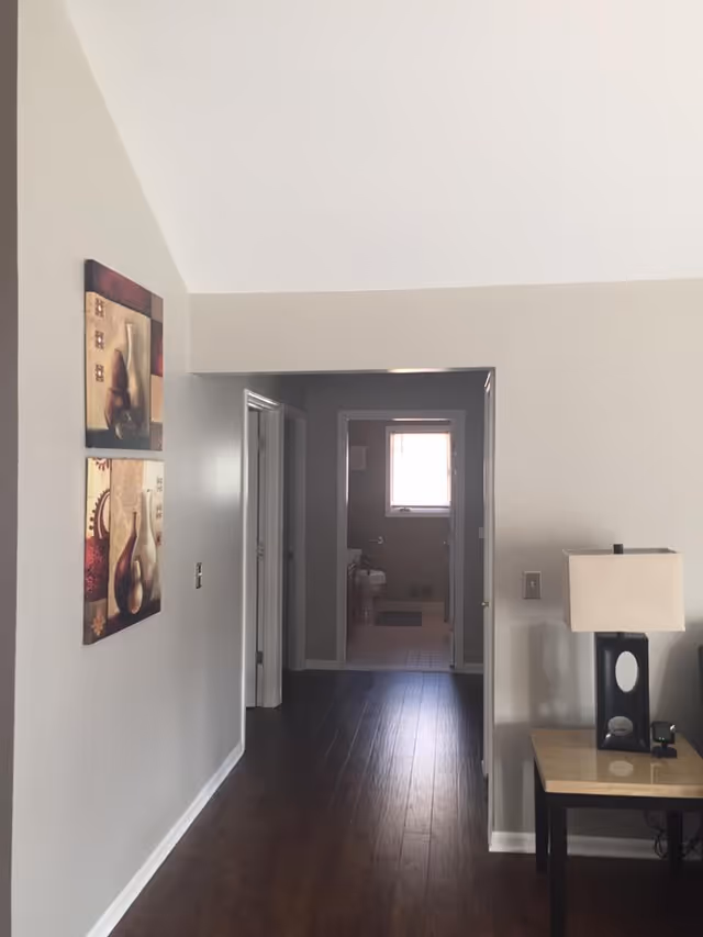 View down a hallway with dark wooden floors leading to a bathroom at the end. The bathroom has a window letting in natural light. On the left wall of the hallway, there are two framed artworks featuring vases. On the right side, there is a small table with a lamp and some electronic devices. The walls are painted light gray.