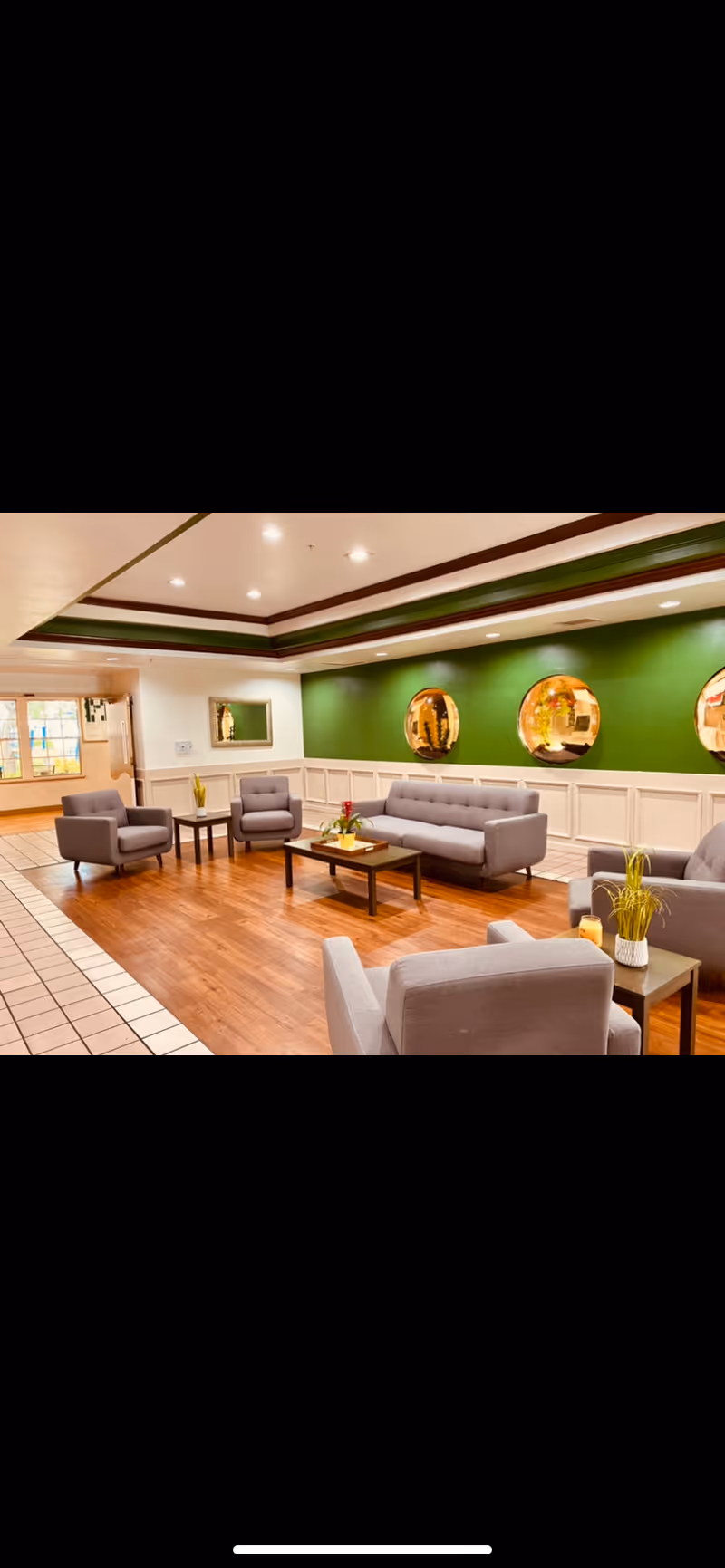 Bright common lounge with gray sofas and armchairs arranged around coffee tables in front of a green accent wall with round mirrors.