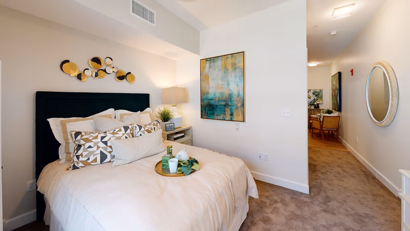 A cozy bedroom with a neatly made bed featuring multiple decorative pillows and a beige comforter. A small nightstand with a lamp, plant, and framed photo is beside the bed. The wall above the bed has circular decorative art, and another abstract painting hangs on the adjacent wall. A hallway with carpet leads to another room with chairs and artwork visible. A round mirror is mounted on the hallway wall.