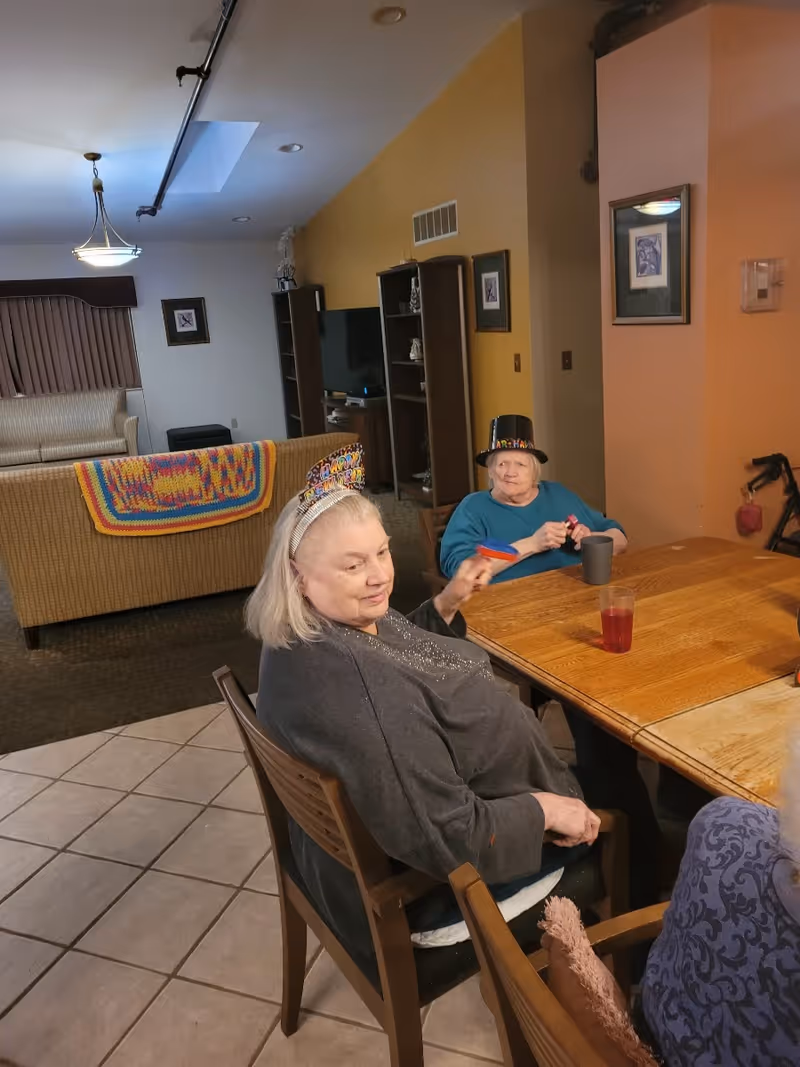 Two elderly women sitting at a wooden table in a living room area. One woman is wearing a colorful birthday hat and a gray sweater, while the other woman is wearing a blue top and a black birthday hat. Behind them is a couch with a colorful crocheted blanket draped over it, a TV, and shelves with decorative items. The room has tiled flooring and warm lighting.