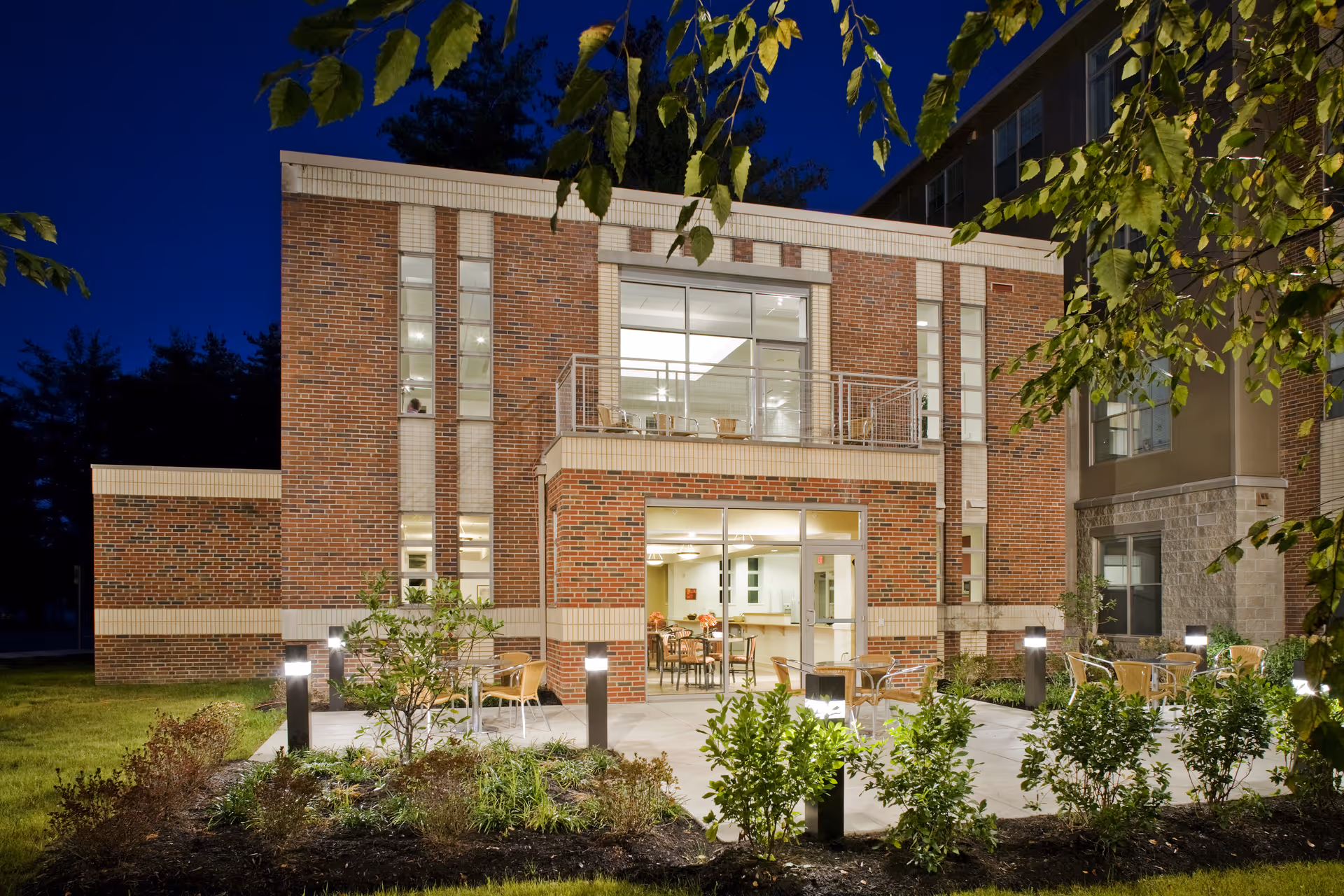 Nighttime exterior view of a brick retirement residence with a lit interior common room, balcony, and patio seating surrounded by landscaping.