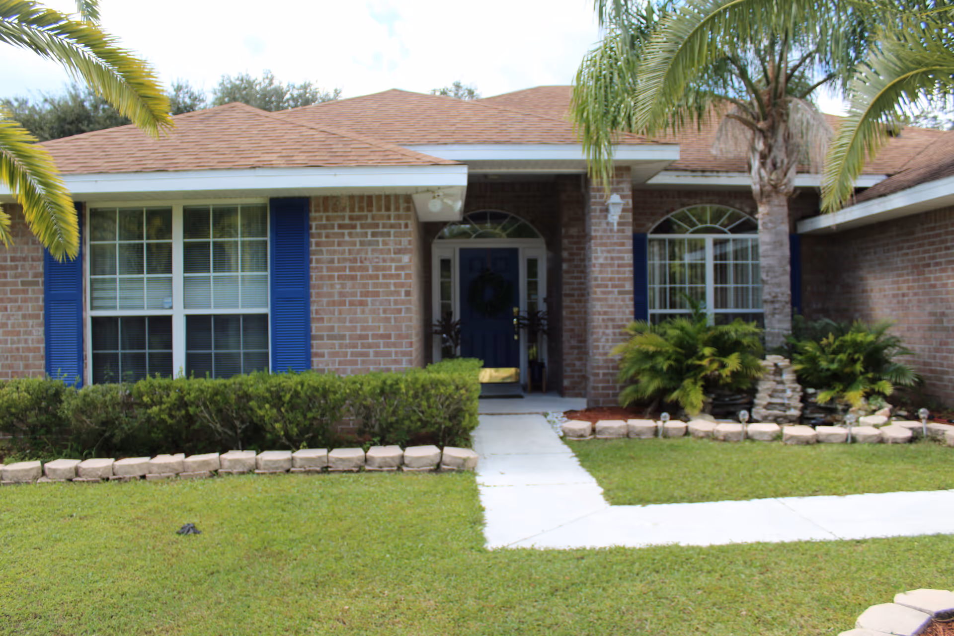 Front exterior view of a single-story brick house with a brown shingled roof, blue shutters on the windows, a blue front door with a wreath, and a well-maintained lawn with bushes and palm trees.