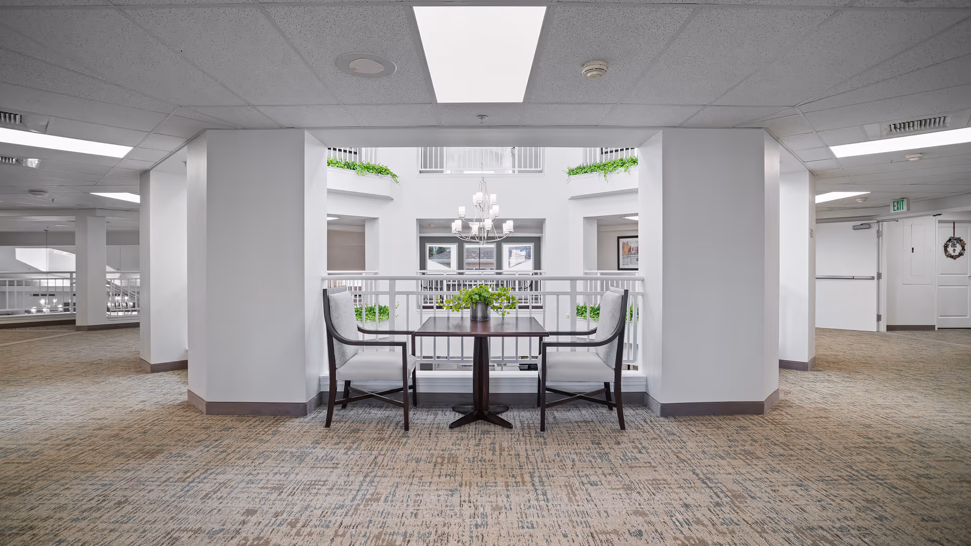 A bright and spacious interior hallway area in a senior living facility with a small table and two chairs positioned near a railing overlooking a lower level. The space features white walls, a patterned carpet, and a large skylight above. Green plants and a chandelier are visible in the open atrium below.