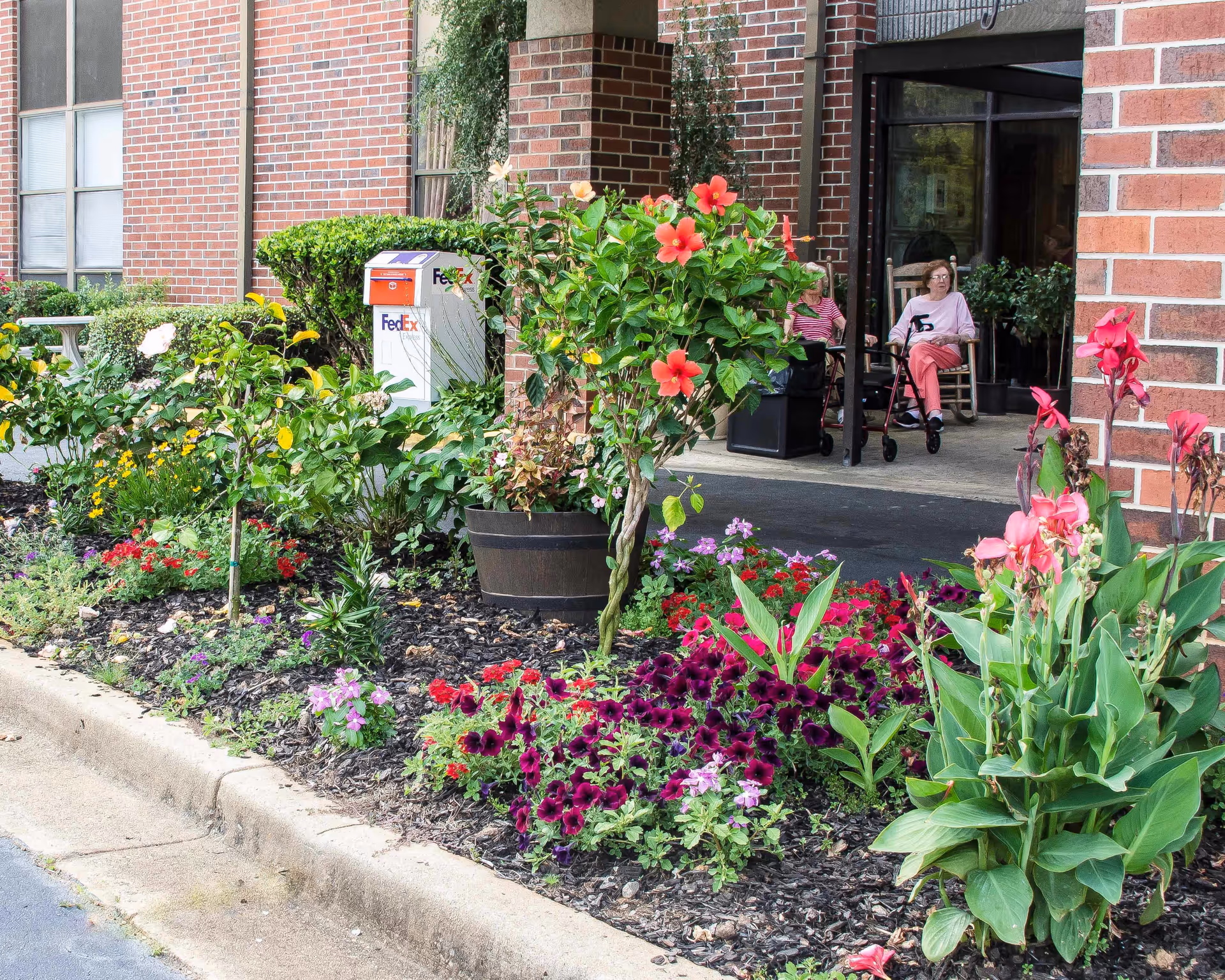A colorful flower garden with various blooming flowers and green plants in front of a brick building entrance. Two elderly women are sitting on rocking chairs near the entrance, one with a walker. A FedEx drop box is visible near the garden.