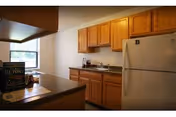 A kitchen area with wooden cabinets, a white refrigerator, a countertop with a granite-like surface, and a window letting in natural light. There are some items on the countertop including a box and a few small objects near the sink.
