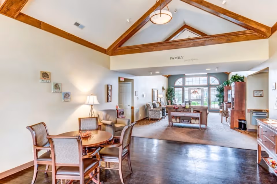 A bright and spacious senior living facility common area with wooden beams on a vaulted ceiling. The room features a round wooden table with four chairs in the foreground, a cozy seating area with armchairs and sofas in the background, large windows letting in natural light, and decorative plants. The word 'FAMILY' is displayed on the wall above the seating area.