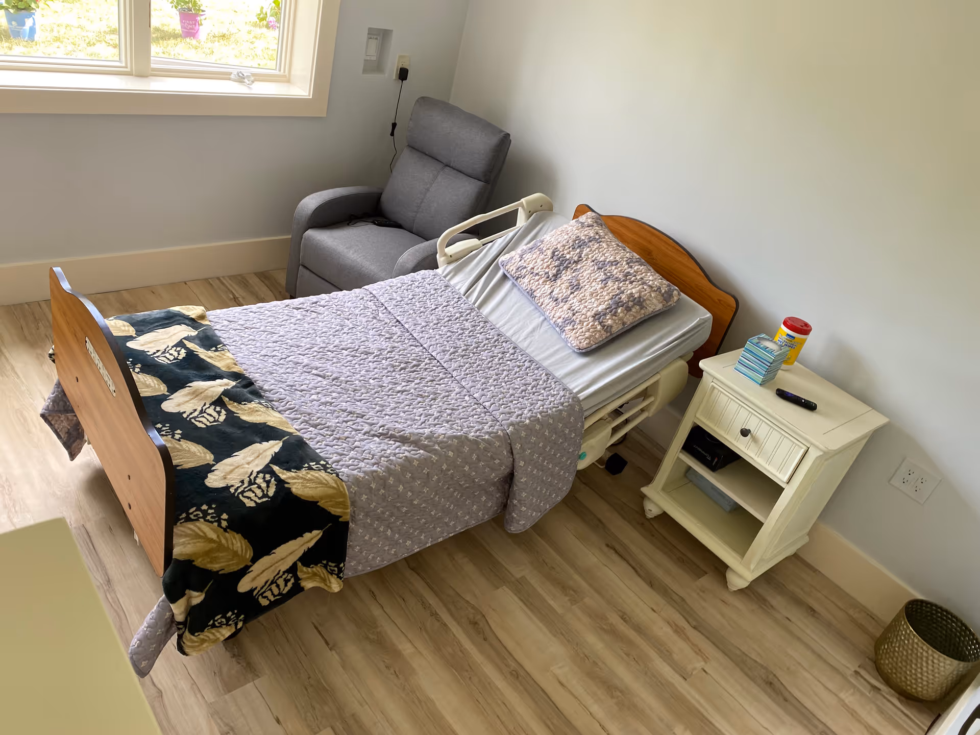 A senior living bedroom with a hospital-style bed covered with a gray quilt and a decorative pillow. Next to the bed is a white nightstand with a remote control, a container of disinfecting wipes, and a stack of tissues. A gray recliner chair is positioned near a window with a view of the outdoors. The room has light wood flooring and white walls.