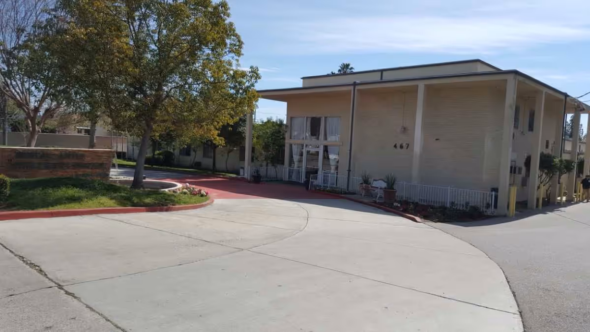 Driveway and front entrance of a two-story beige medical convalescent hospital with trees and landscaping.