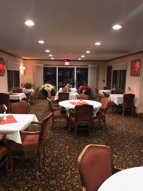 Interior view of a dining room with multiple round and square tables covered with white tablecloths and red placemats. Each table is surrounded by upholstered chairs with wooden frames. The room has patterned carpet flooring, beige walls, and recessed ceiling lights. There are windows on the side walls and a glass door at the far end with an exit sign above it. Potted plants and floral decorations are placed near the windows and door.