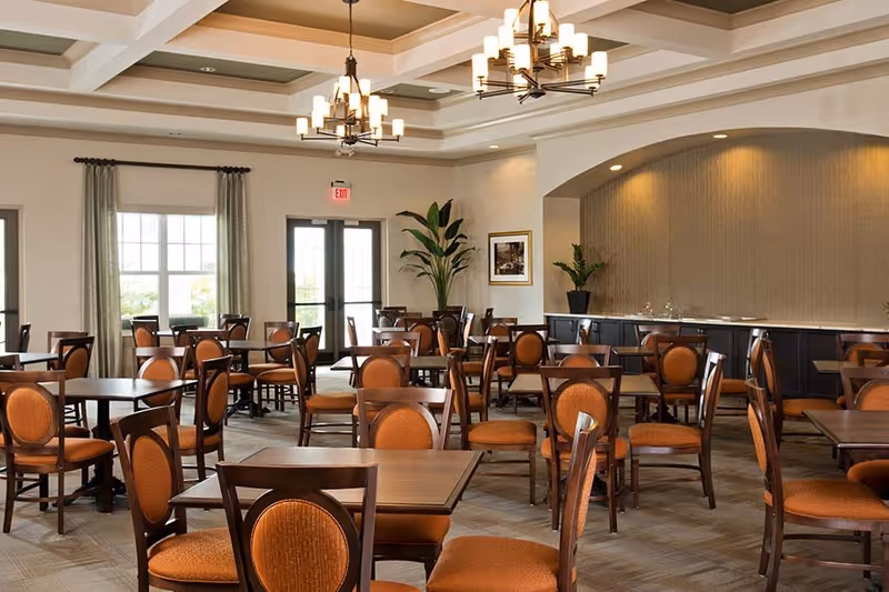 A spacious dining room with multiple wooden tables and orange cushioned chairs arranged neatly. The room features large windows with curtains, two chandeliers hanging from a coffered ceiling, and a sideboard with decorative plants and framed artwork on the wall.