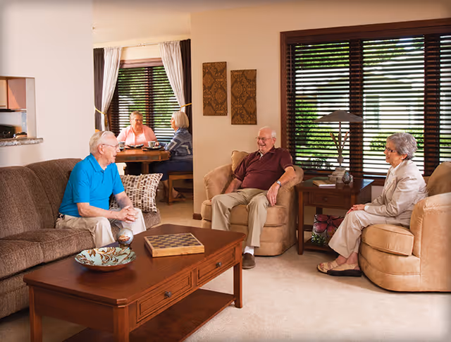 A cozy living room in a senior living facility with three elderly people sitting and chatting on comfortable chairs and a sofa. In the background, two more elderly individuals are seated at a dining table near a window with curtains. The room is warmly lit with wooden furniture and decorative wall art.