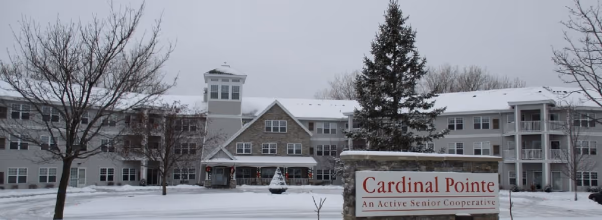 Exterior view of Cardinal Pointe of Maplewood senior living facility during winter, showing a large multi-story building with snow-covered roof and ground, leafless trees, and a sign in front that reads 'Cardinal Pointe An Active Senior Cooperative'.