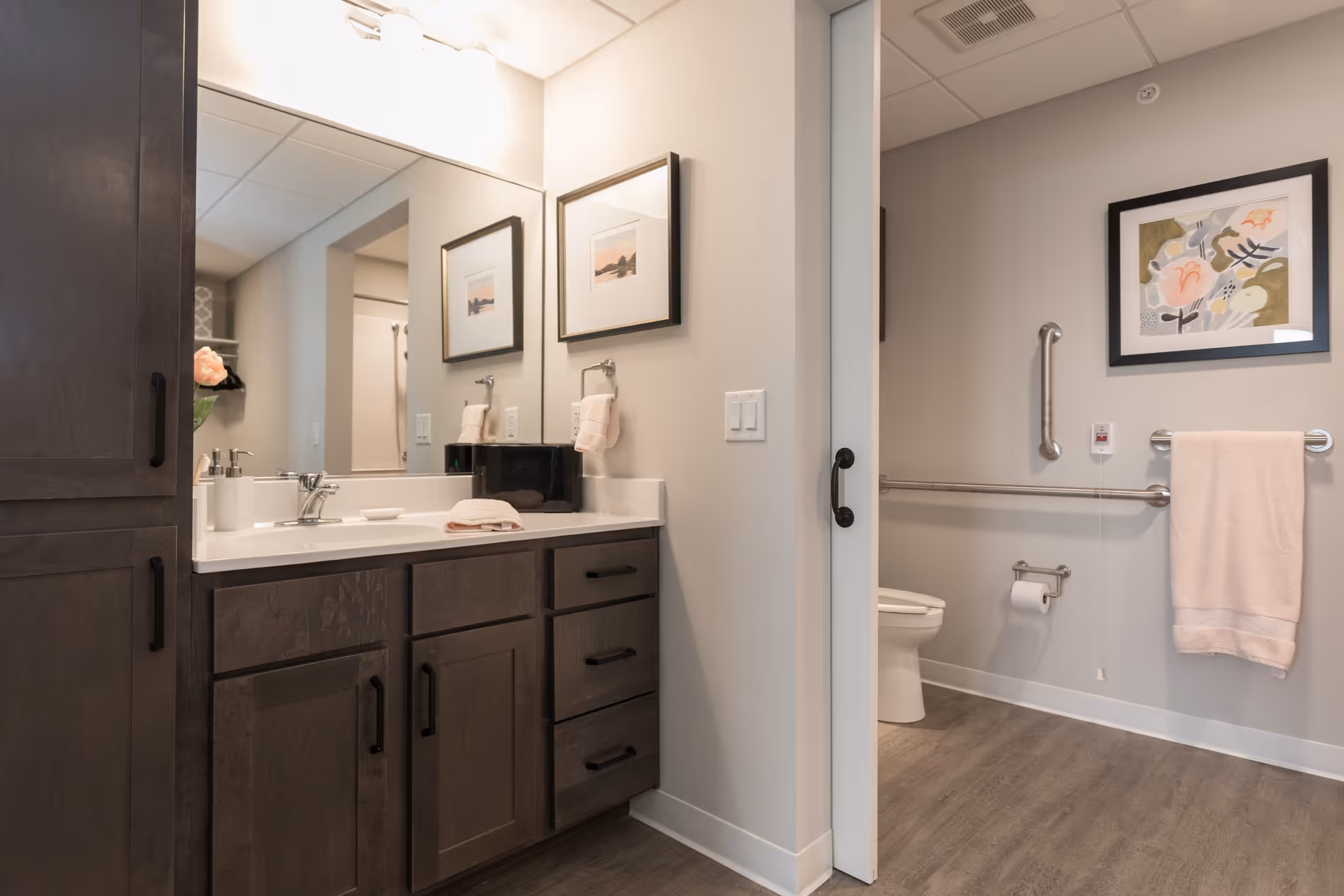 A clean and modern bathroom in a senior living facility featuring a dark wood vanity with a white countertop and sink, a large mirror above the sink, and a partially open door revealing a toilet area with grab bars, a towel rack with a pink towel, and framed artwork on the wall.