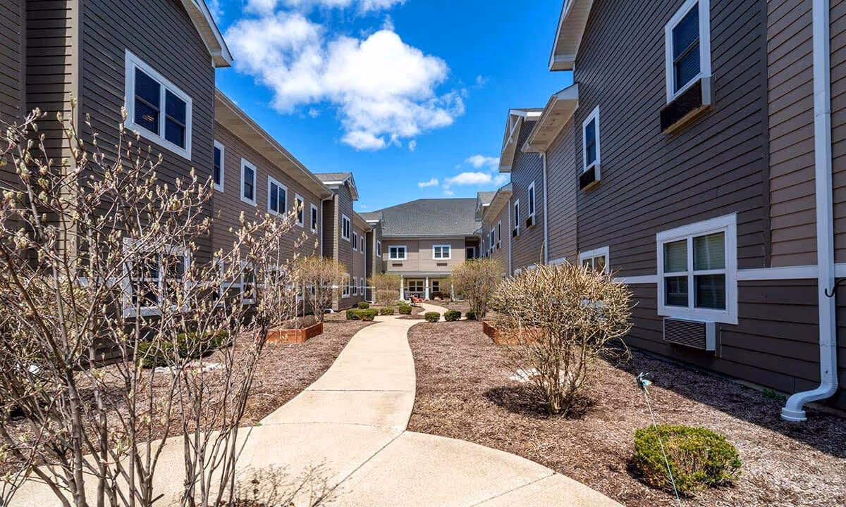 Curved courtyard walkway between two brown multi-story residential buildings with shrubs and a blue sky.
