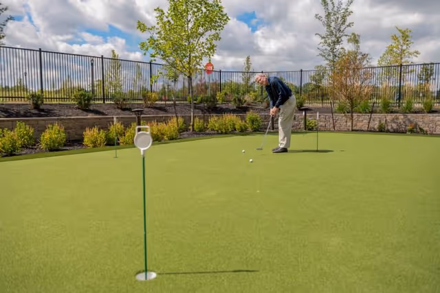 A man practicing putting on an outdoor putting green with flags and landscaped beds under a partly cloudy sky.