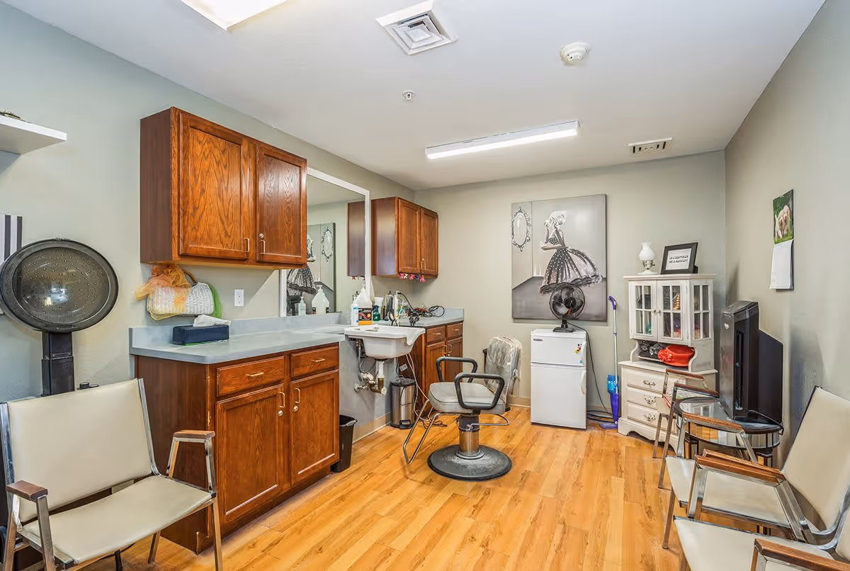Interior of a small salon or beauty room with wooden cabinets, a countertop with a sink, a salon chair, a hair dryer, and several chairs arranged around the room. The room has light-colored walls, wood flooring, a small refrigerator, a fan, a TV, and decorative artwork on the walls.