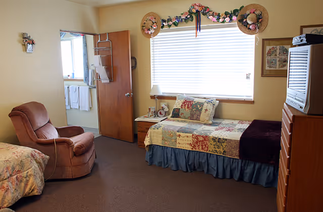 A cozy bedroom with a single bed covered in a patchwork quilt, a cushioned armchair, a wooden dresser with a TV on top, and a window decorated with floral garlands and hats. The door is open, revealing a bathroom with towels hanging on racks.