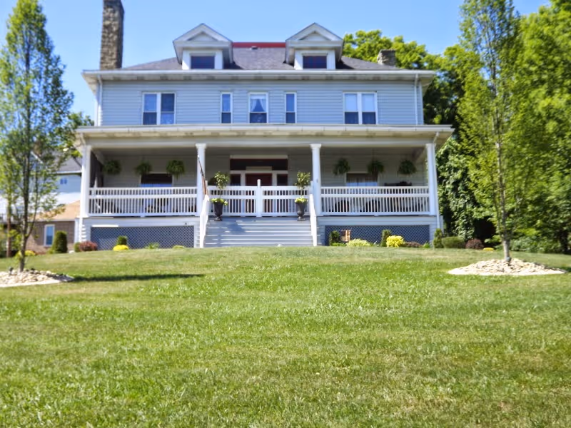 Front exterior view of a large two-story house with a wide porch, white railing, and several windows. The house is surrounded by a well-maintained green lawn with small trees and shrubs on either side. The sky is clear and blue.