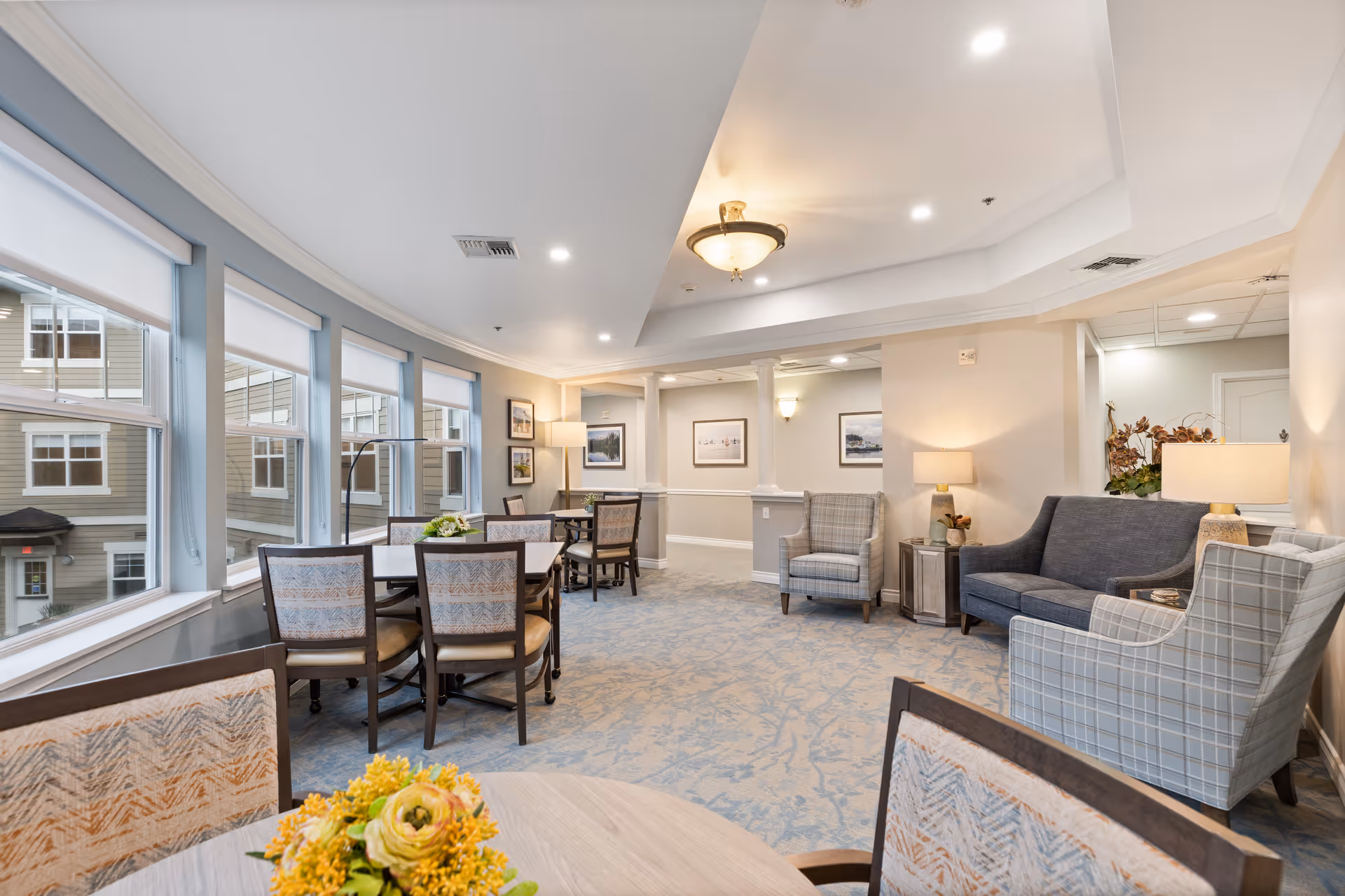 A bright and spacious common area in a senior living facility featuring multiple tables with chairs near large windows, comfortable armchairs, a sofa, and soft lighting from lamps and ceiling fixtures. The room has light-colored walls and carpeted floors with a subtle pattern.
