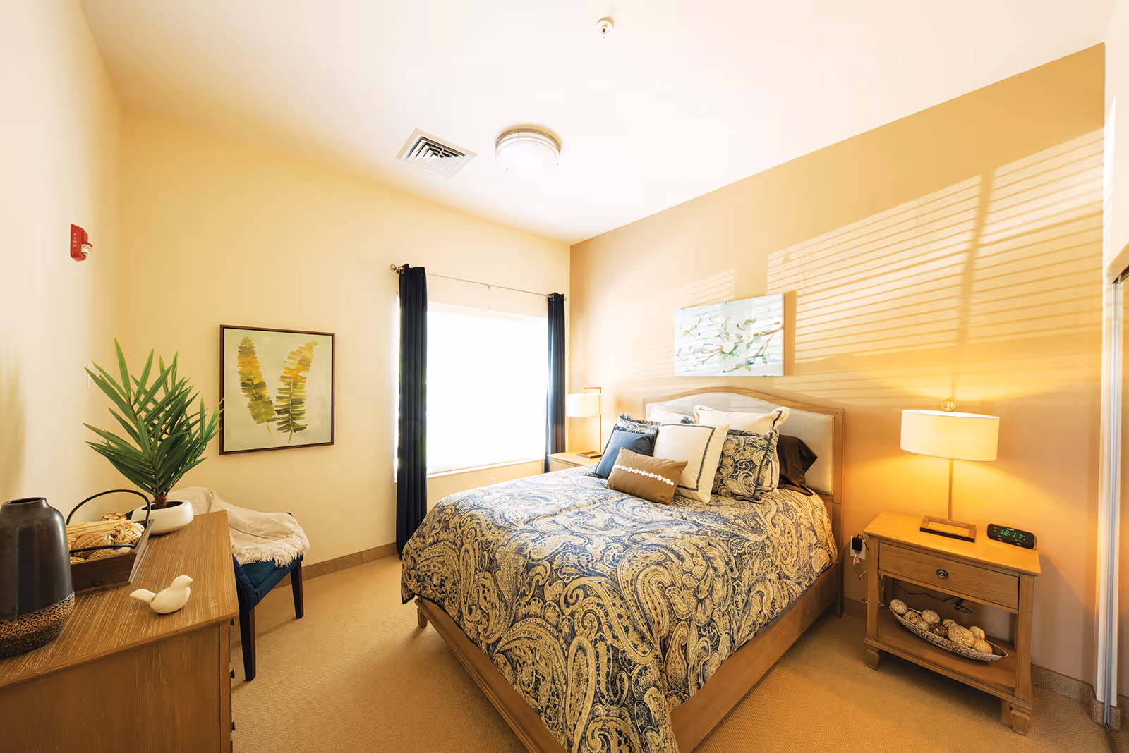 A cozy bedroom with a large window covered by dark curtains, a bed with a patterned blue and beige comforter and multiple pillows, a wooden nightstand with a lamp and clock, a wooden dresser with decorative items including a plant and a small bird figurine, and a framed artwork on the wall.