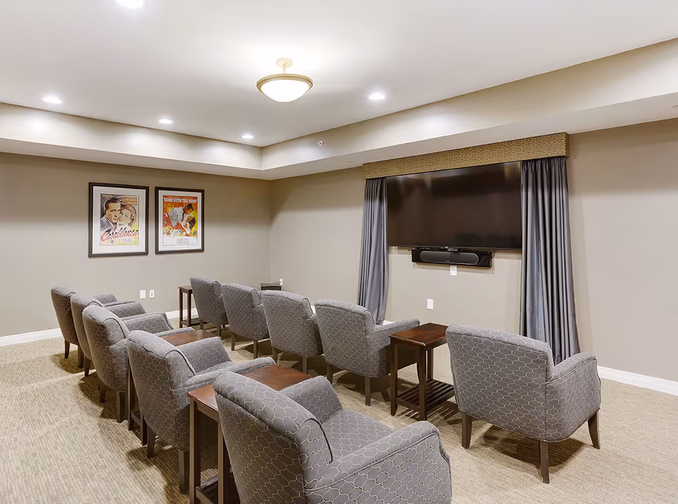A small theater room with two rows of gray patterned armchairs facing a large flat-screen TV mounted on the wall. The room has beige walls, carpeted floor, recessed lighting, and two framed vintage movie posters on one wall.