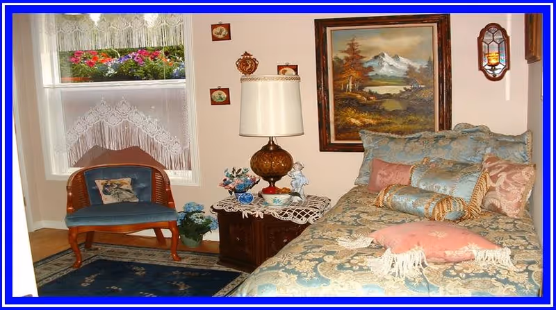 A cozy bedroom with a bed covered in a blue and gold patterned bedspread and multiple pillows. Next to the bed is a wooden nightstand with a decorative lamp, a small figurine, and a vase with flowers. A wooden chair with a blue cushion and a pillow is placed near a window with lace curtains and a flower box outside. The walls are decorated with framed paintings and small wall hangings.