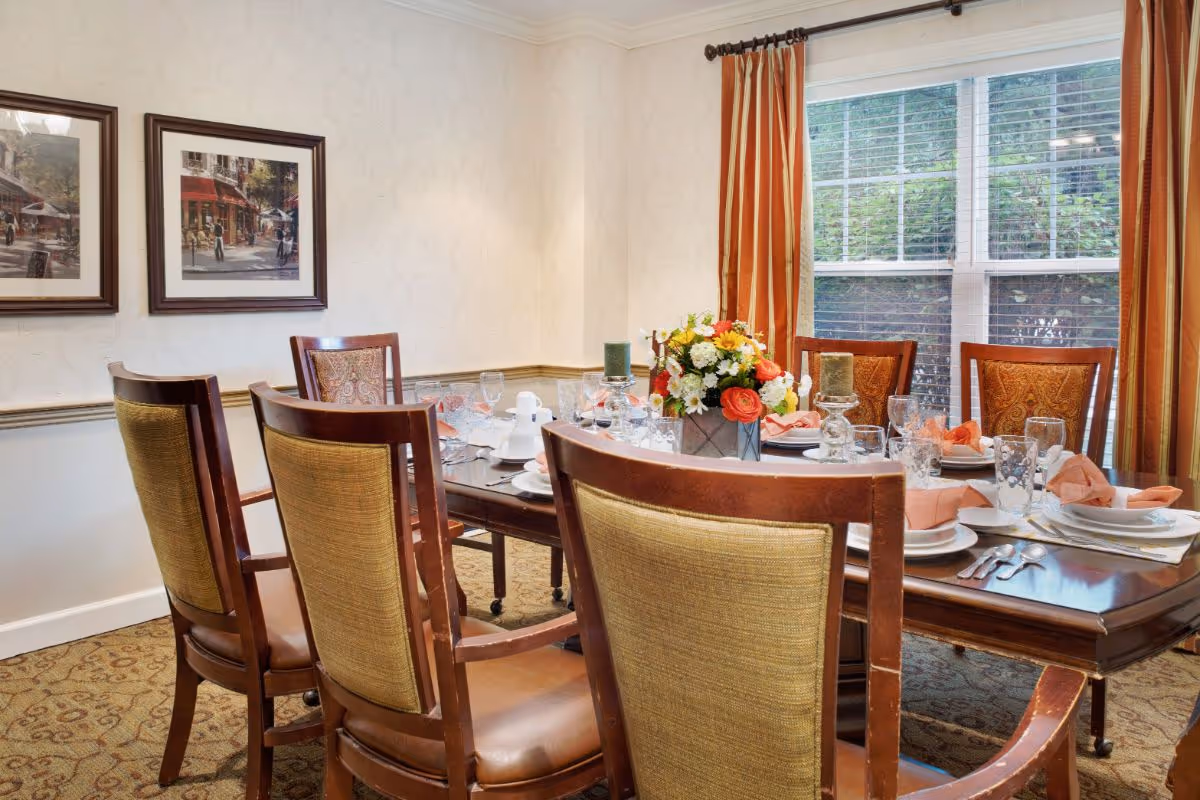 A formal dining room with a set table, wooden chairs, a floral centerpiece and a large window with curtains.