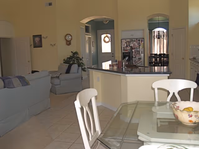 Interior view of a residential living space showing a glass dining table with white chairs in the foreground, a kitchen area with a counter and sink in the midground, and a living room with sofas and a wall clock in the background. The walls are painted yellow and there are arched doorways leading to other rooms.