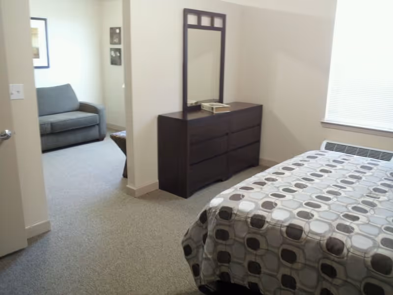 A furnished bedroom with a patterned bedspread, dark dresser with mirror, and adjacent seating nook.