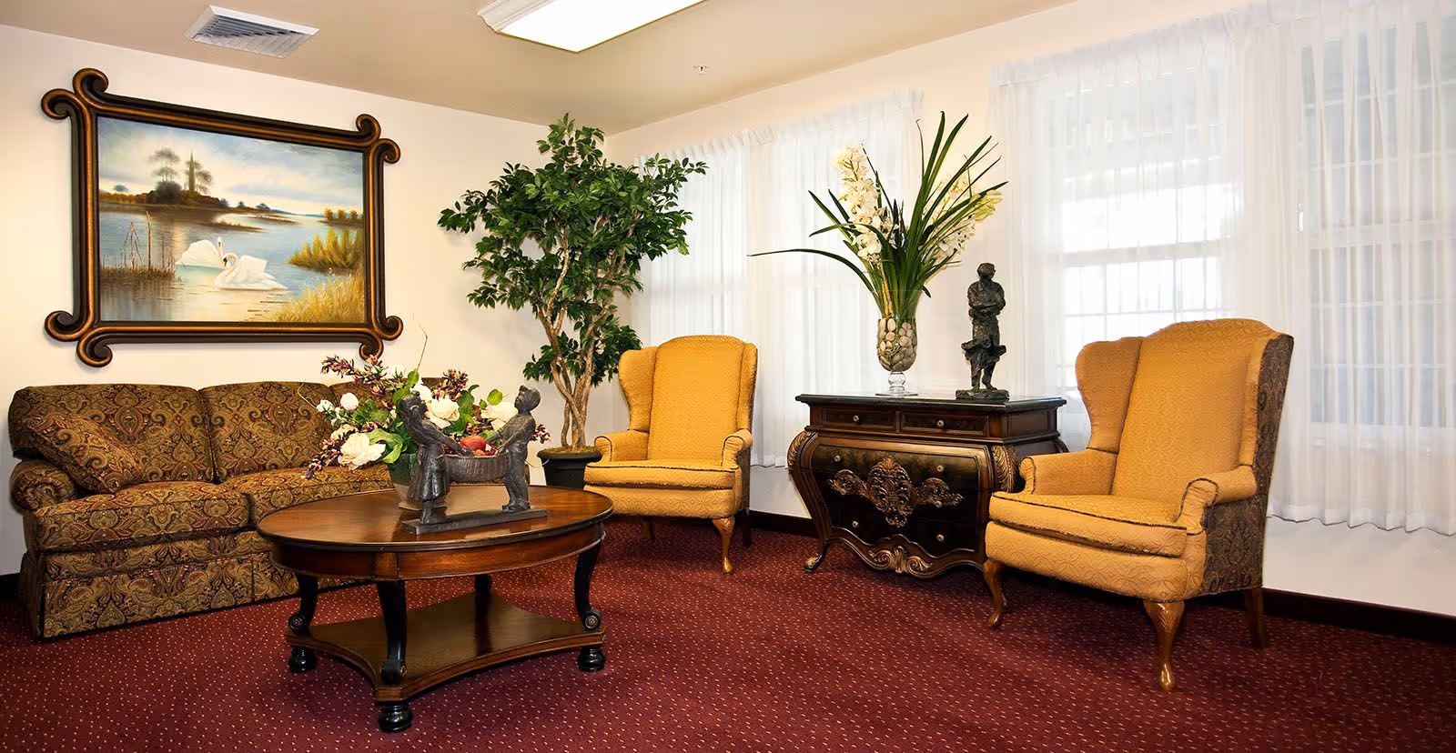A cozy living room with a patterned sofa, two mustard yellow wingback chairs, a wooden coffee table with a floral centerpiece, a decorative chest with a vase of flowers and a small statue, a large potted plant, and a framed painting of swans on a lake hanging on the wall.