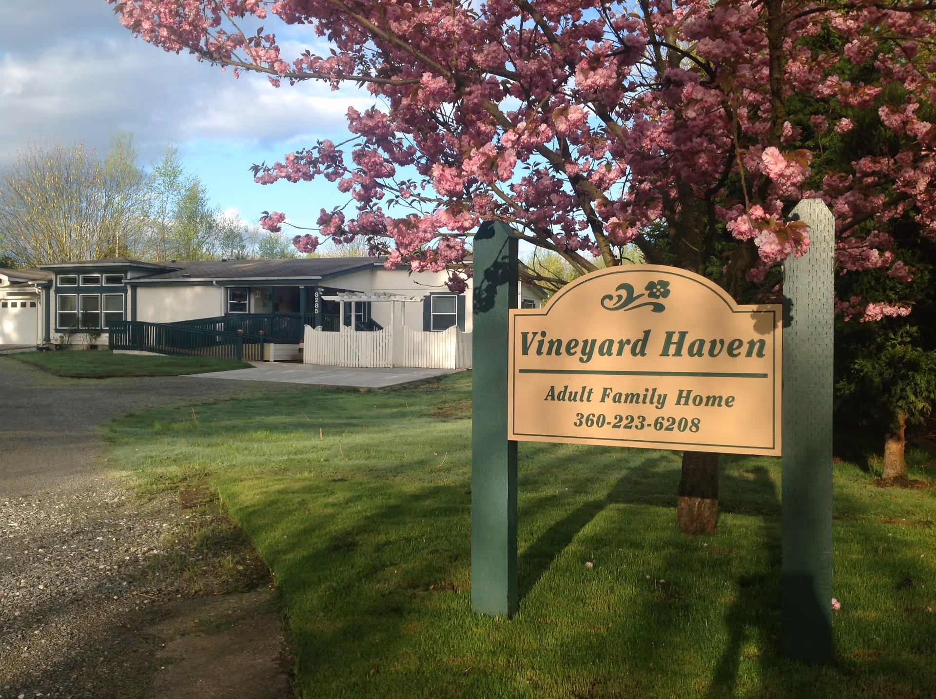 A wooden sign for Vineyard Haven Adult Family Home stands on a lawn in front of a single-story residential building with a pink blossoming tree.