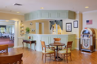 Interior view of a senior living facility common area with a light green reception desk, a round wooden table with four chairs, a vintage jukebox, and a small table with decorative items. The space has wooden flooring and light yellow walls, with an American flag hanging on the wall.