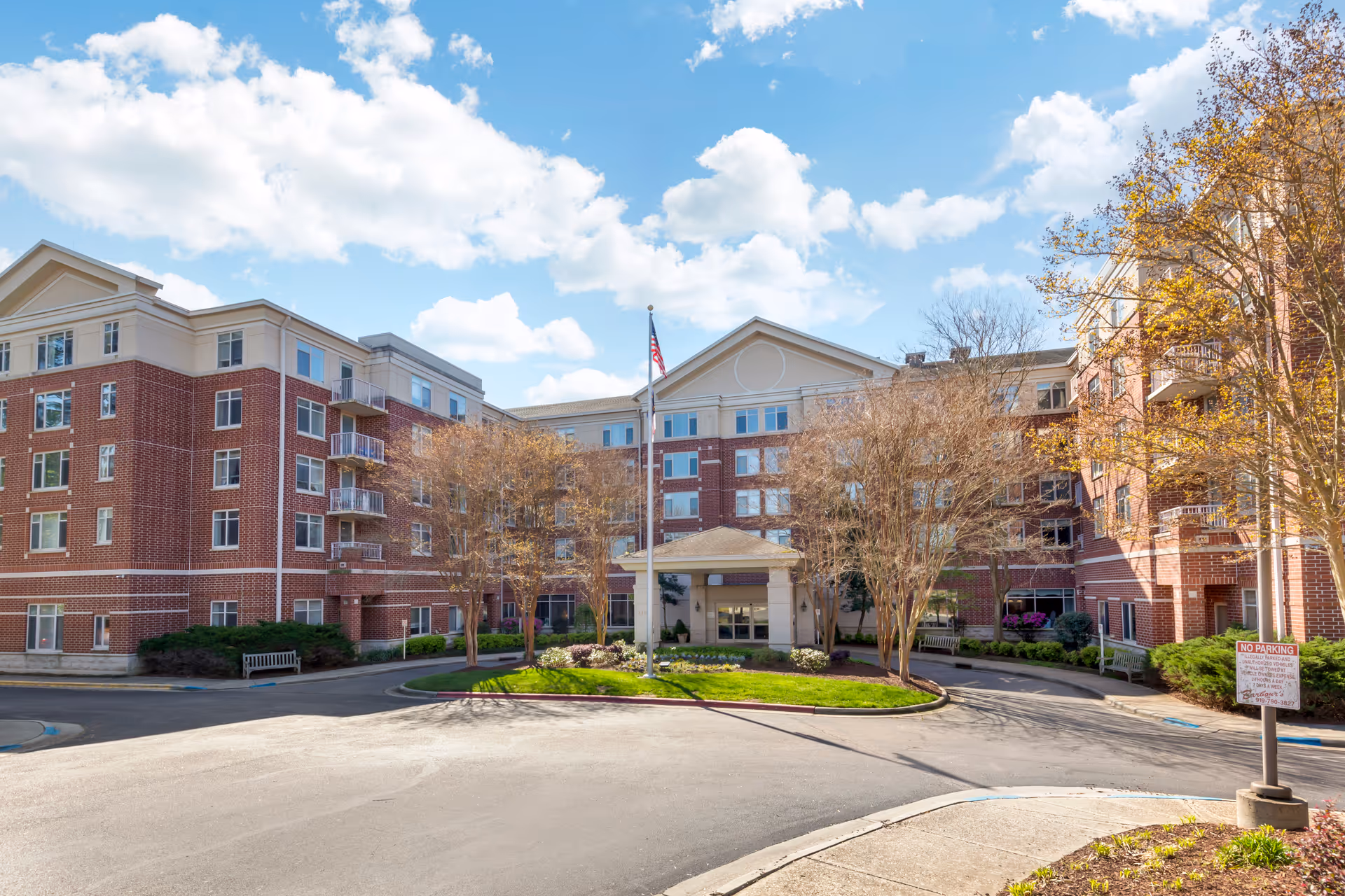 Front exterior view of a multi-story senior living facility building with red brick and beige accents, a circular driveway, landscaped greenery, and an American flag on a flagpole under a partly cloudy blue sky.