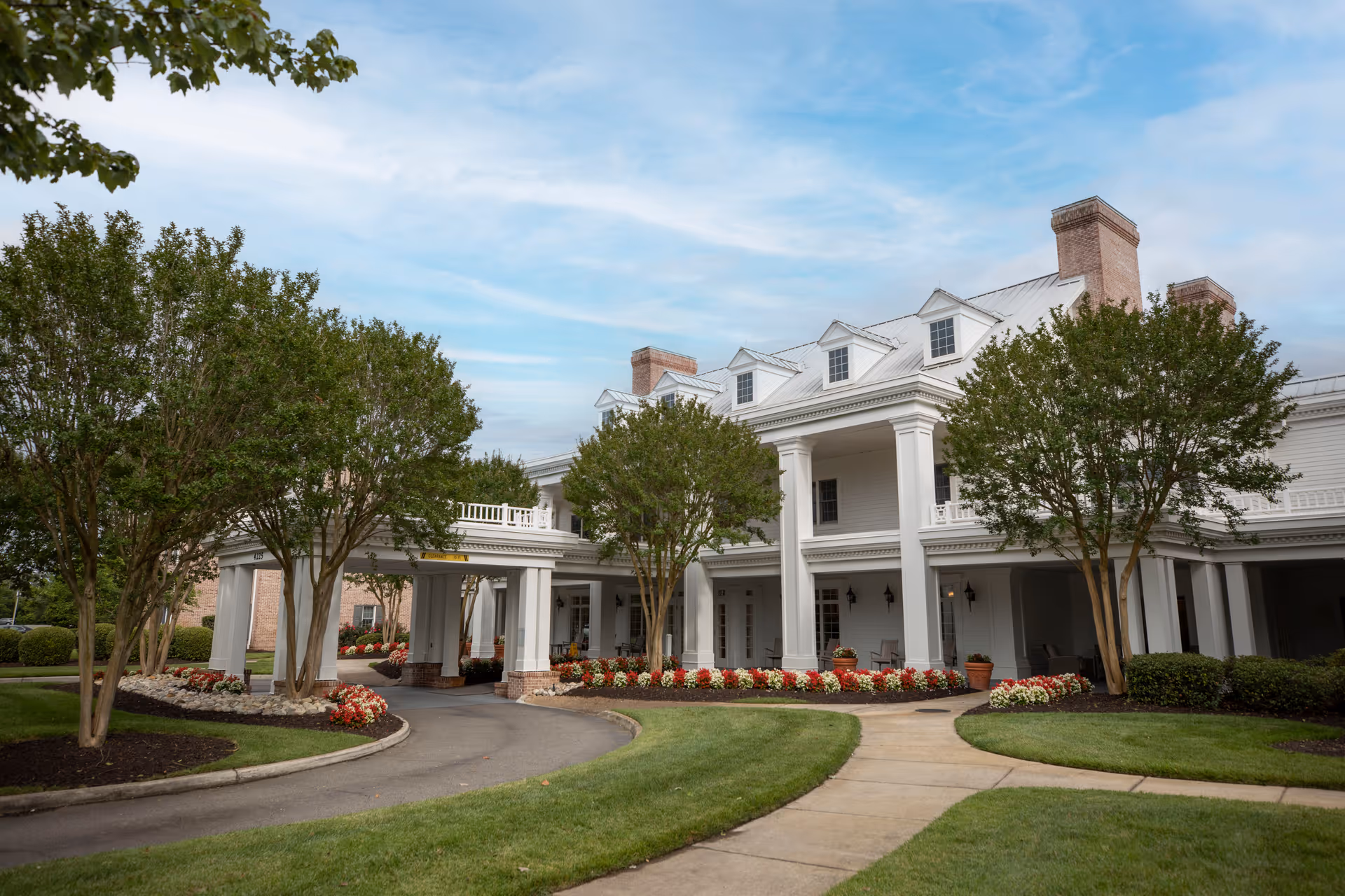 Front entrance of a white, columned retirement community building with a circular driveway, landscaped flower beds, and trees.