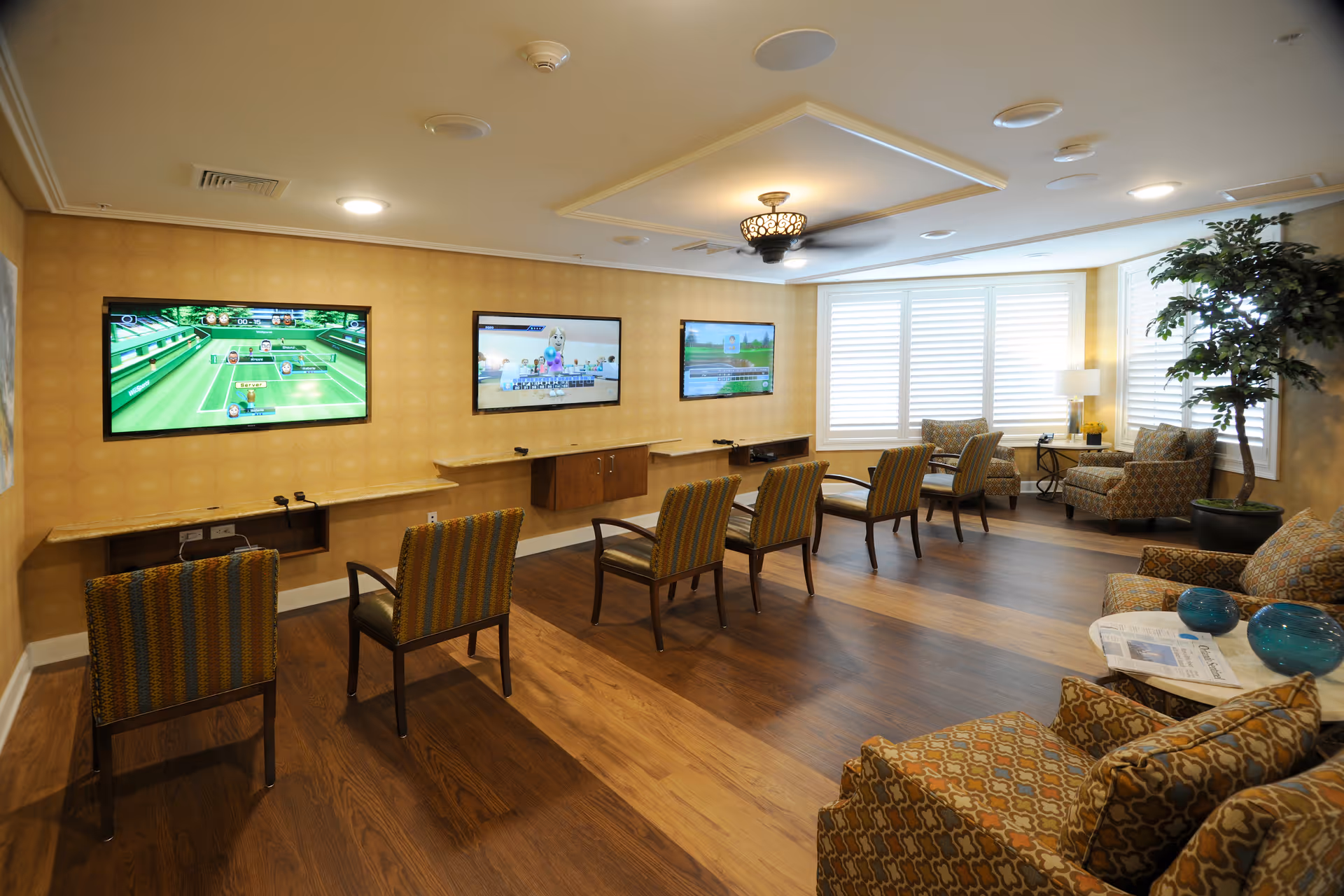A spacious room with three wall-mounted televisions displaying video games, several chairs arranged in front of the screens, and comfortable armchairs with patterned upholstery near a window with white blinds. The room has warm yellow walls, wood flooring, and a decorative ceiling light fixture.
