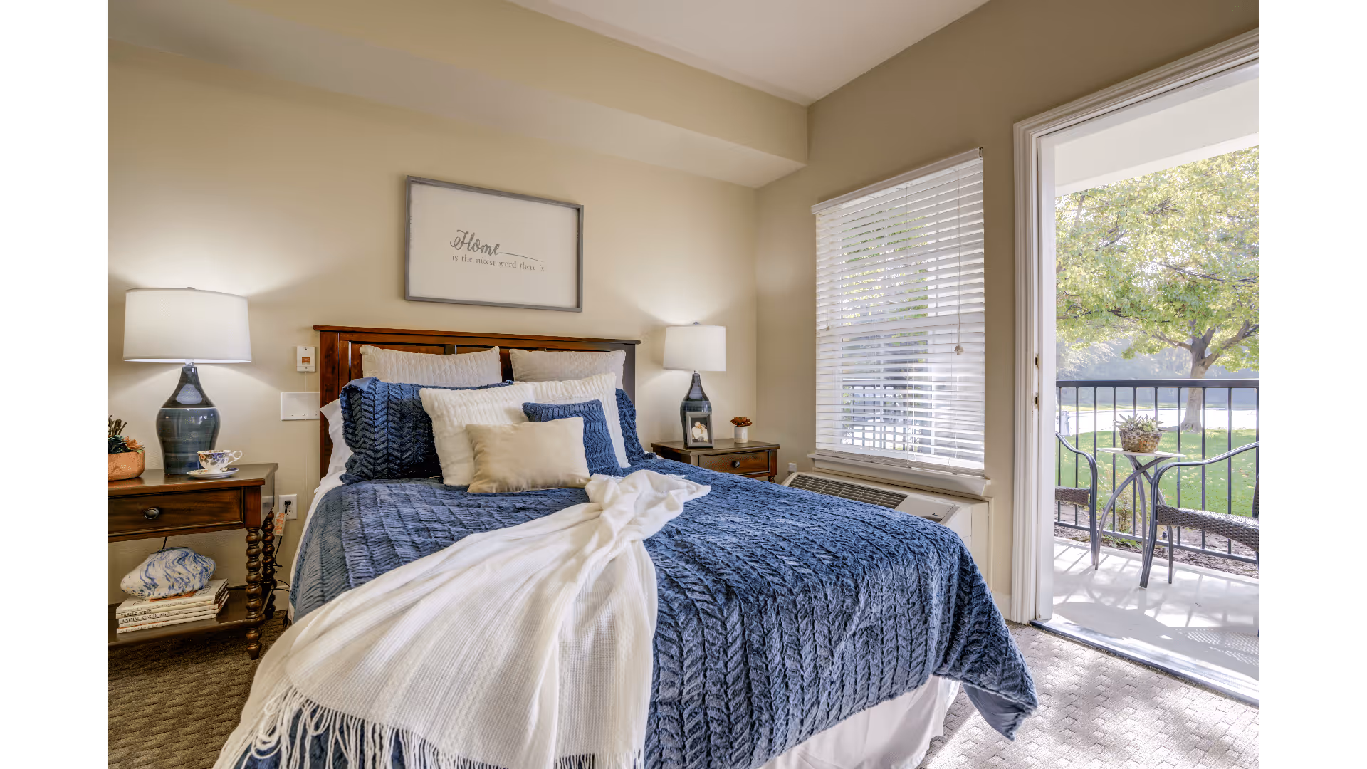 A cozy bedroom with a neatly made bed featuring a blue textured blanket and multiple pillows. Two bedside tables each hold a lamp, and one has a teacup and a small plant. A framed sign above the bed reads 'Home is the nicest word there is.' A window with white blinds and a door open to a balcony with a small table and two chairs overlooking a green outdoor area with trees.