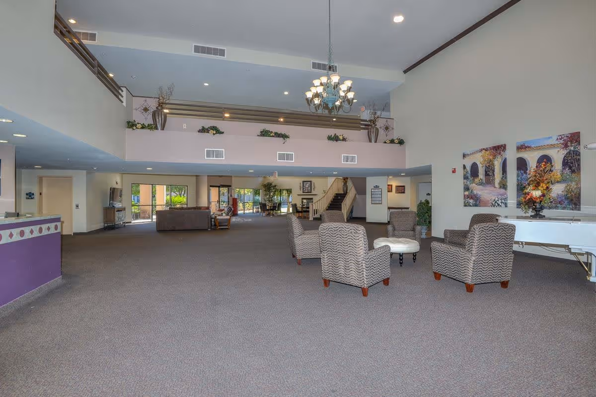 Spacious assisted living facility common area with high ceilings, a chandelier, patterned armchairs arranged around a small round ottoman, a white grand piano, and colorful paintings on the wall. There is a staircase leading to an upper level with decorative plants and vases, and large windows letting in natural light.