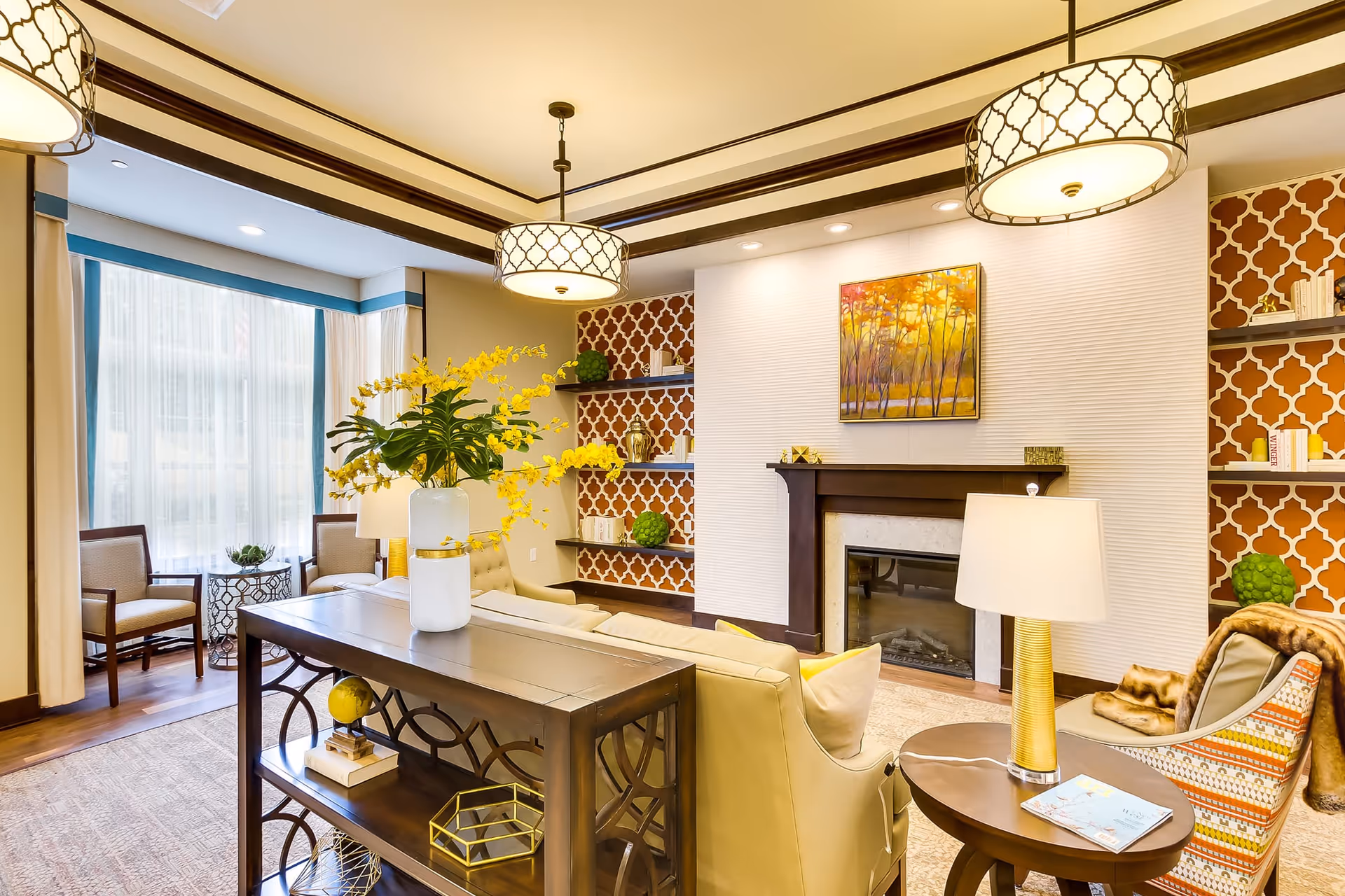 A bright and elegant living room with a beige sofa, a wooden console table with a white vase holding yellow flowers, two armchairs near a large window with sheer curtains, a fireplace with a colorful painting above it, and built-in shelves with decorative items and books. The room features warm lighting from ceiling fixtures and a table lamp.