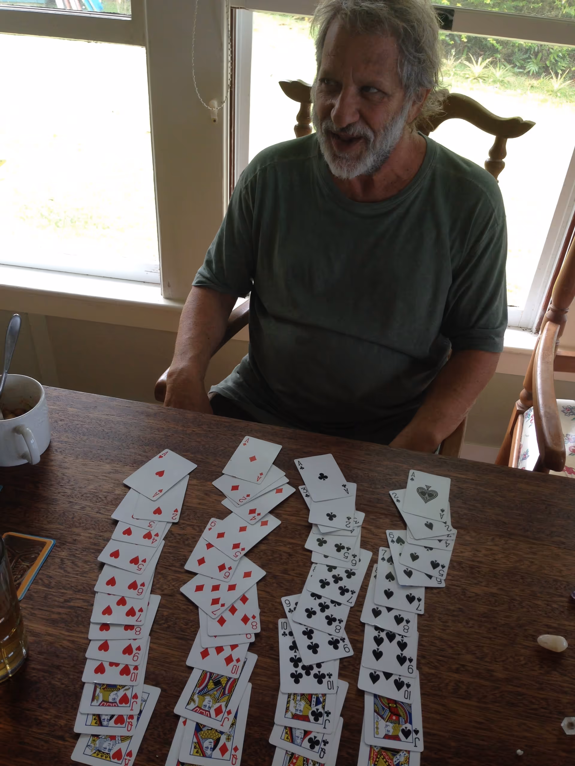An elderly man with gray hair and beard sitting at a wooden table near a window. On the table in front of him are four neat rows of playing cards arranged by suit. There is also a white mug and a glass with a drink on the table.