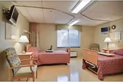A rehabilitation and nursing center room with two hospital beds covered in pink bedspreads, a window with curtains, two chairs, two bedside lamps, and two wall-mounted televisions.